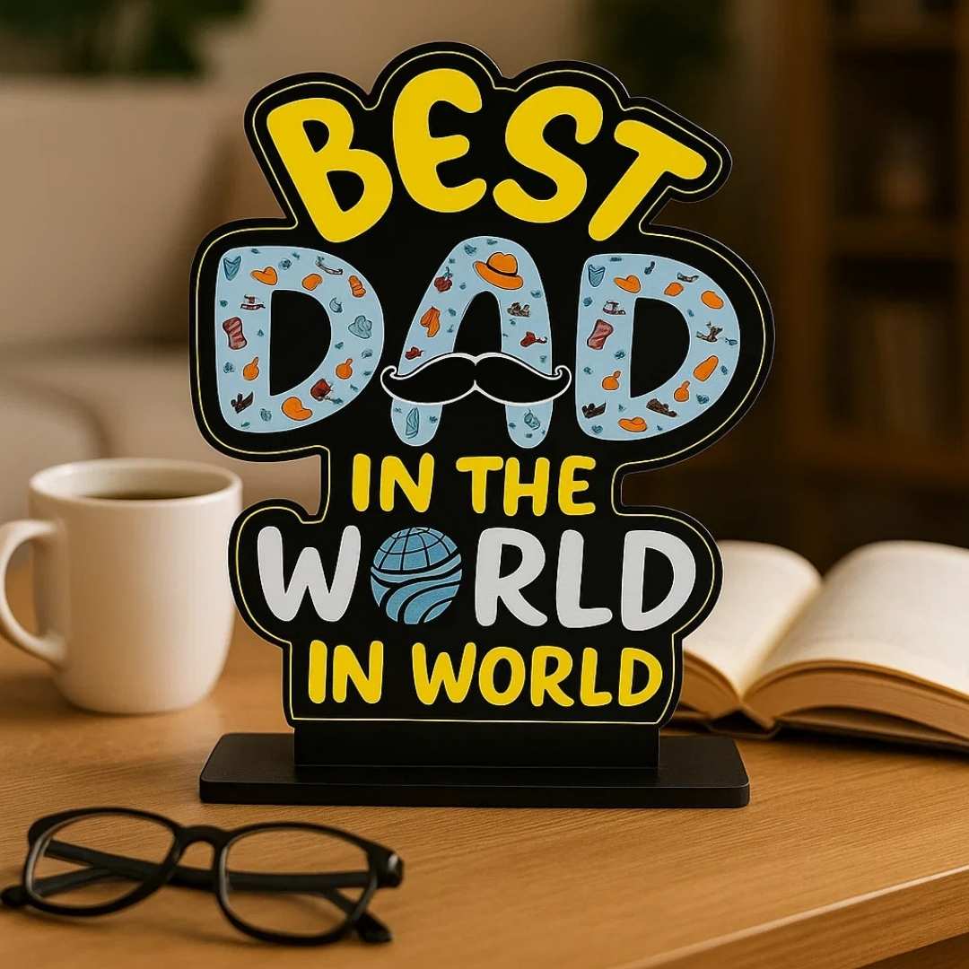 Decorative sign with 'Best Dad in the World' text on a table with a cup, glasses, and book.