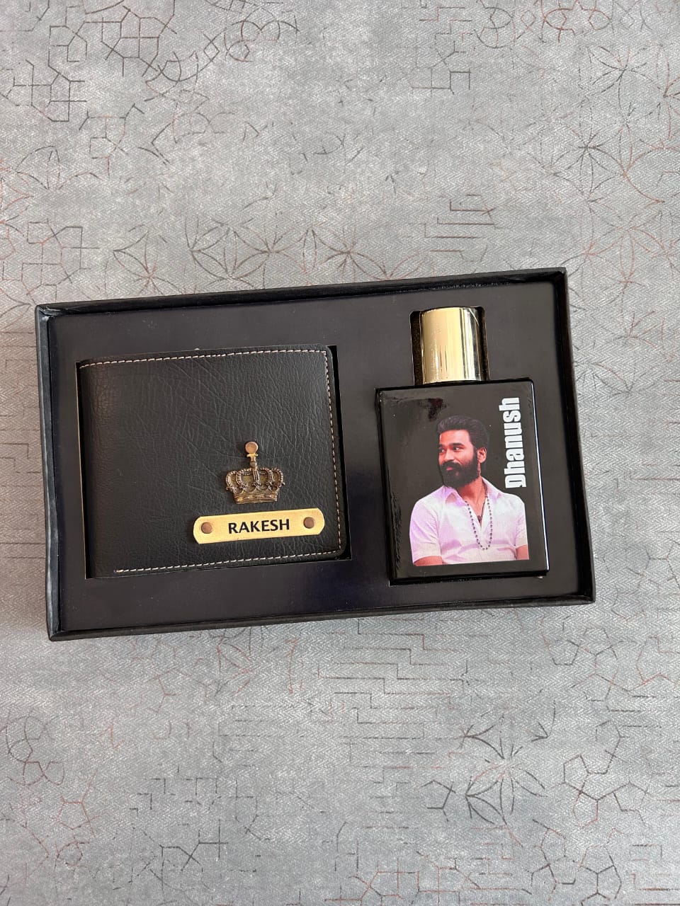 Black wallet and perfume set with 'Rakesh' branding on a textured gray surface