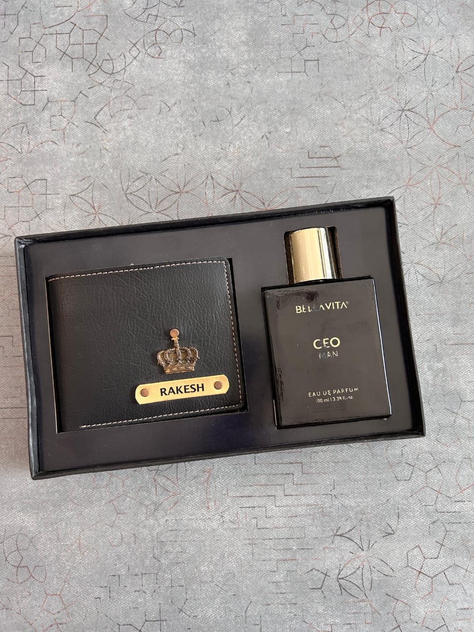 Black leather wallet and perfume set with 'Bella Vita' branding on a textured gray surface