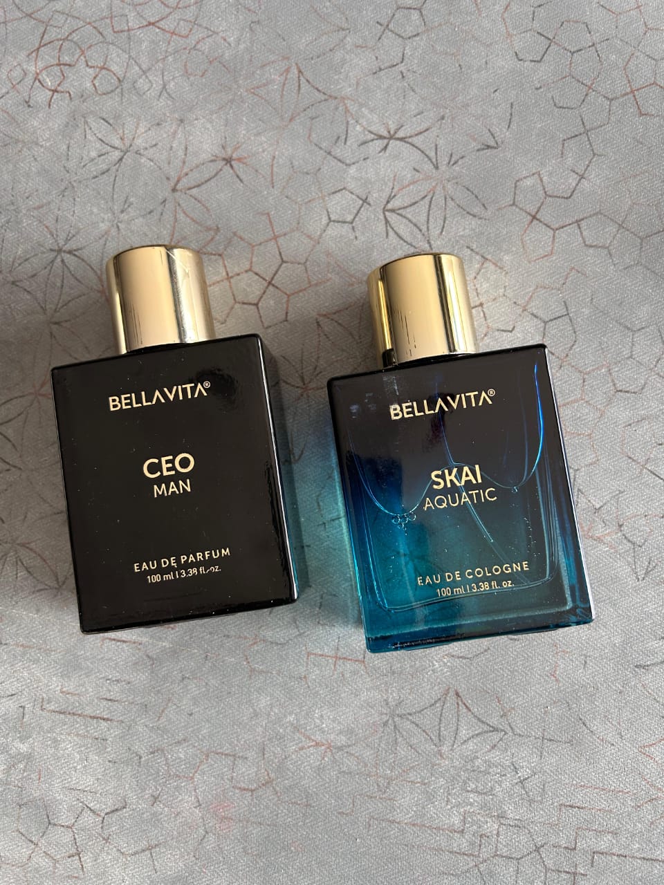 Two Bellavita perfume bottles, one black and one blue, on a textured gray surface.