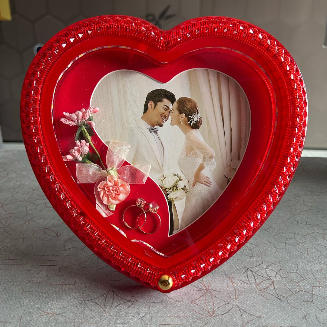 A heart-shaped LED photo frame with a red border displaying a photo of a couple and decorative flowers.