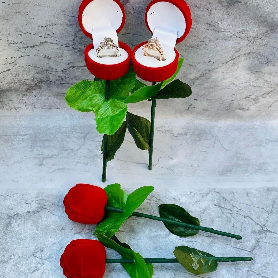 A set of two red rings with fake red flowers, resembling a ring rose, displayed on a marble surface. One ring has a visible engraved message 'I Love You'.