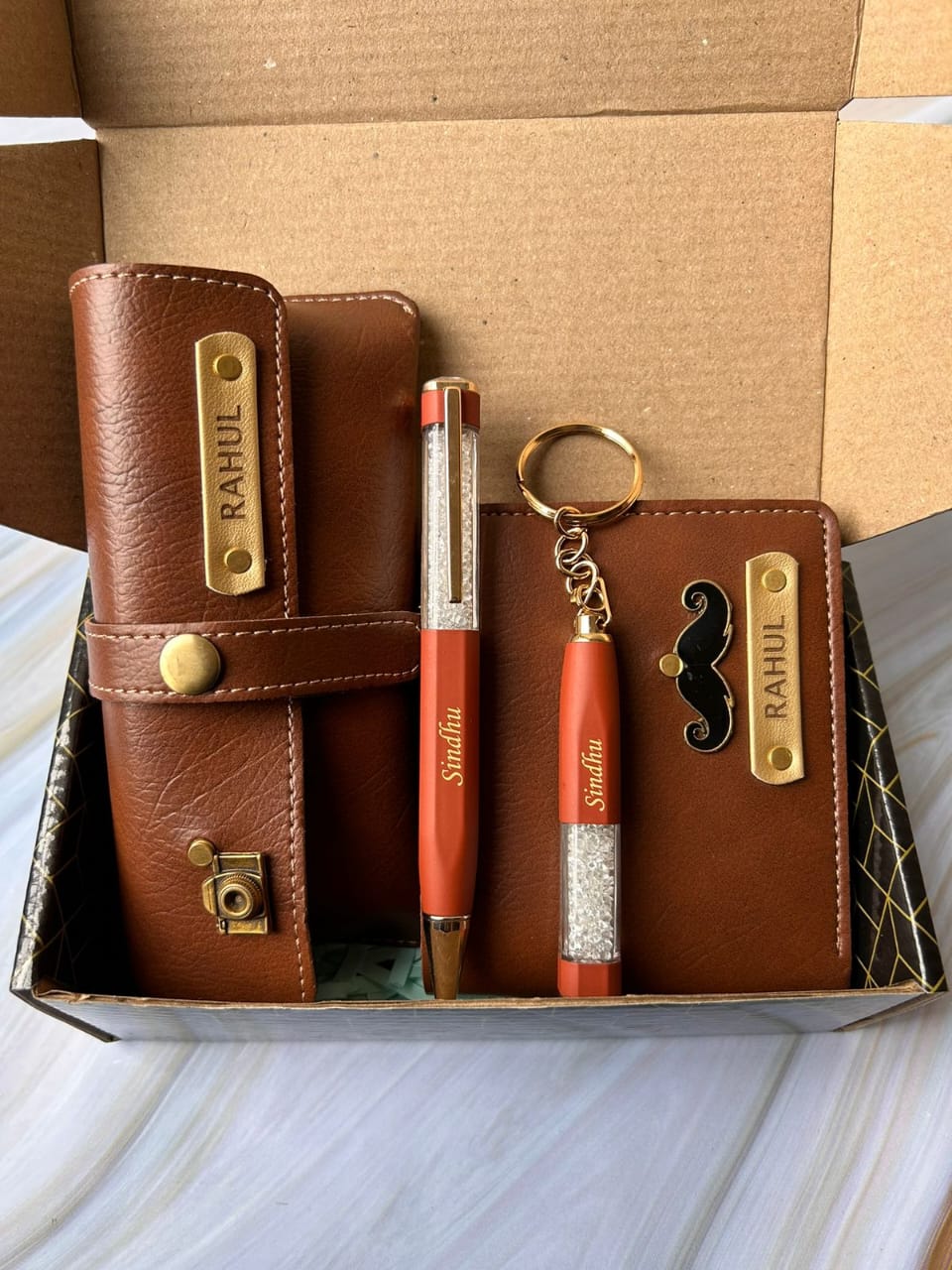 Brown leather wallet, red pen, and keychain with 'Rahul' branding in a box.