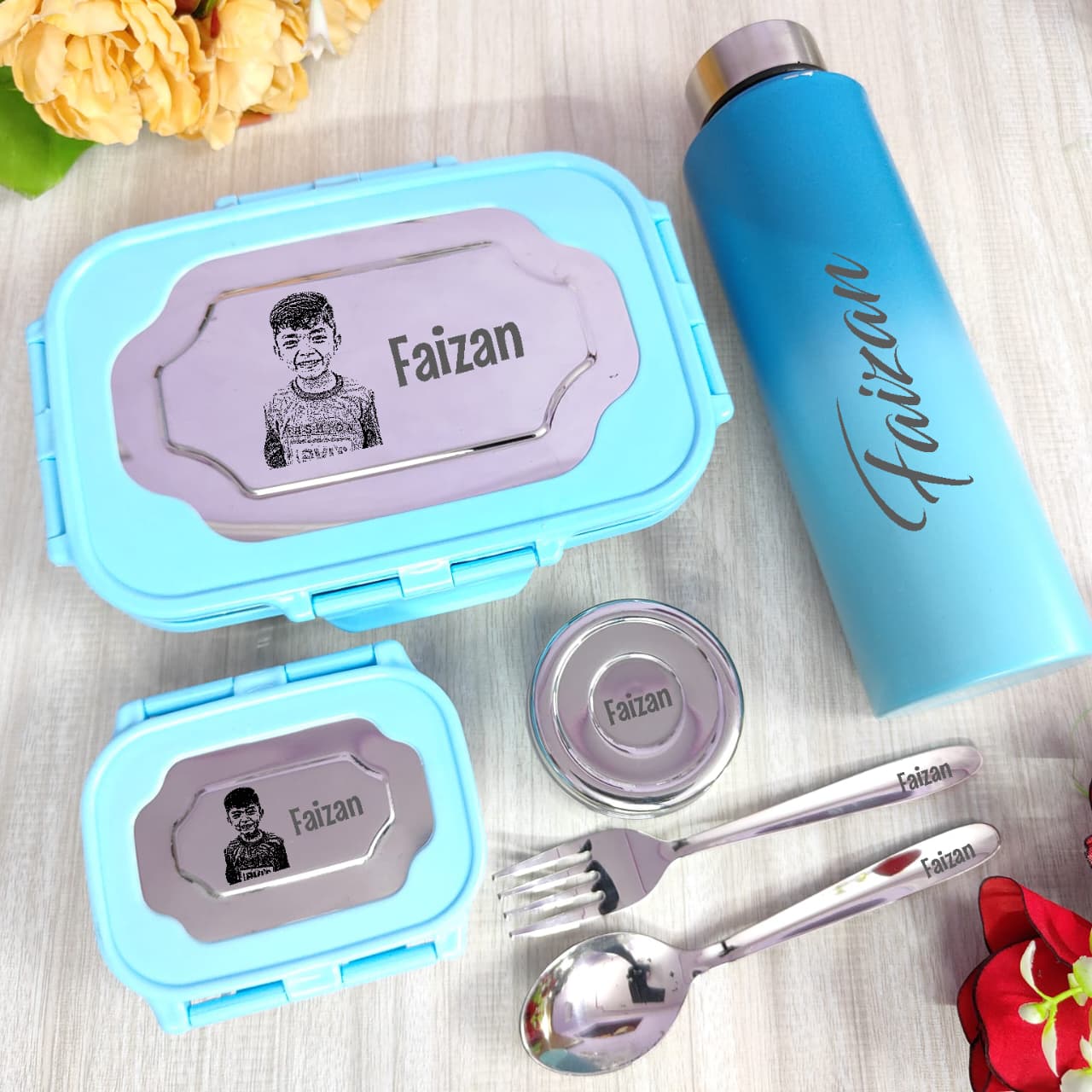 Personalized blue lunch box, bottle, and cutlery set with 'Faizan' branding on a wooden surface.