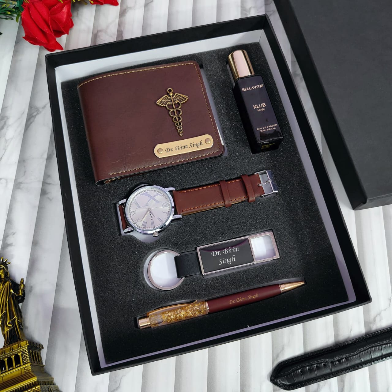 Set of luxury items including a wallet, watch, pen, and keychain in a black box on a marble surface.