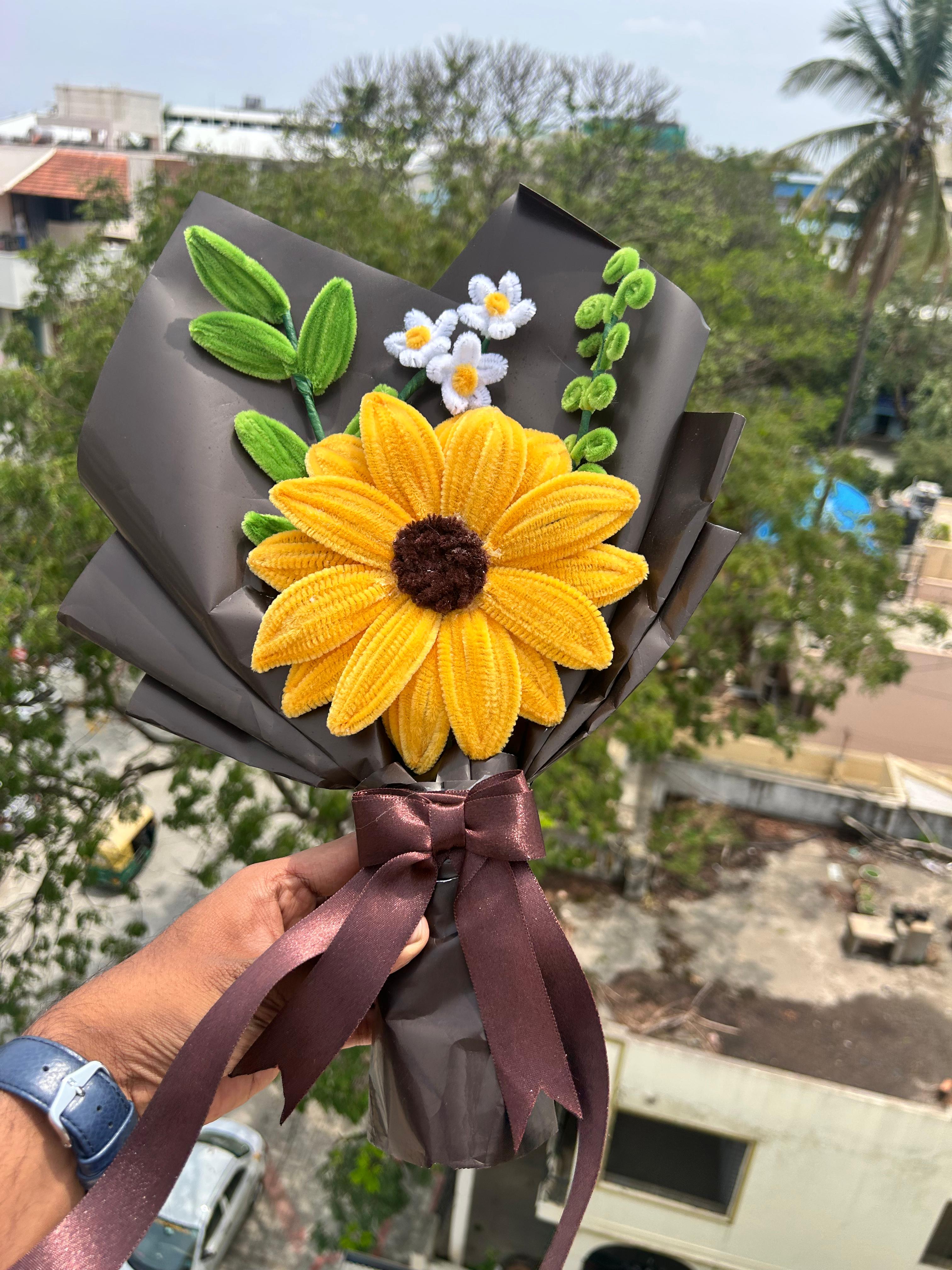 Bouquet with a large yellow sunflower and smaller white flowers, held against an outdoor background.