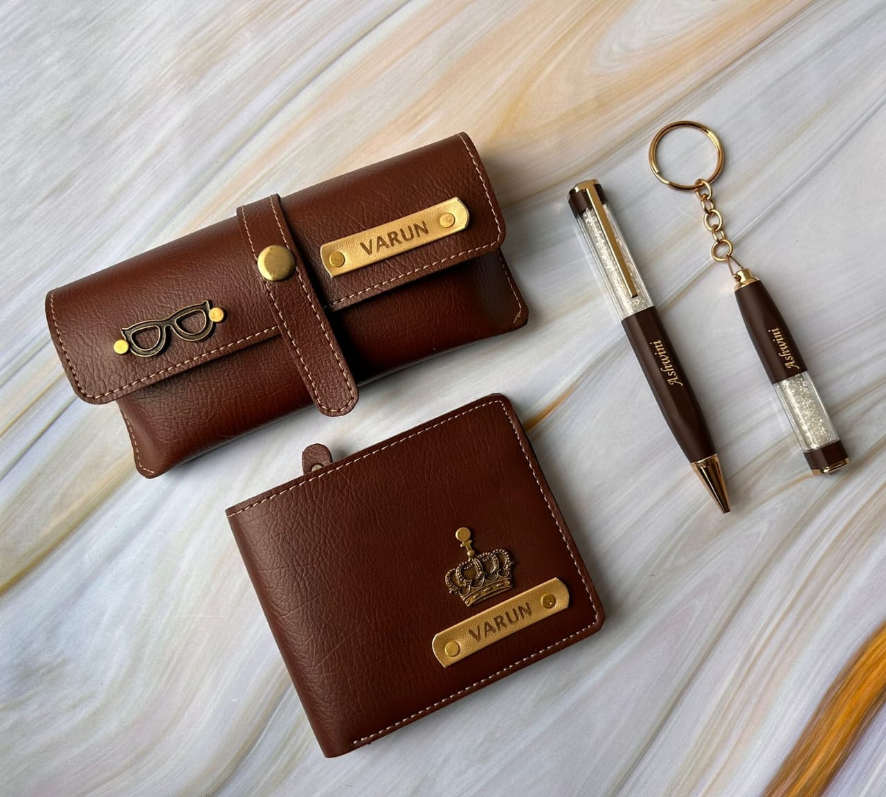Brown leather wallet, pen, and keychain with 'VARUN' branding on a light wooden surface.