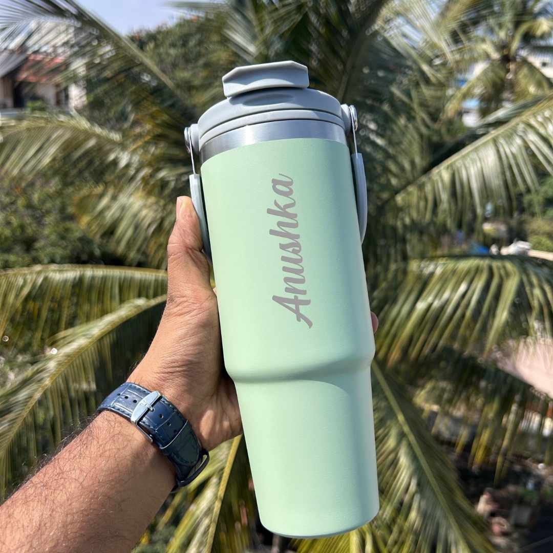 A green personalized flask with the name 'Anushka' engraved on it, being held by a hand with a watch, against a background of palm trees.