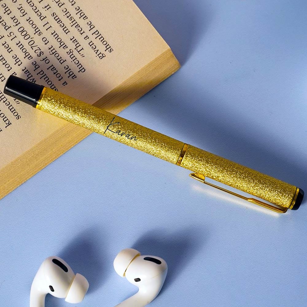 Gold pen with black cap on a blue surface next to white earbuds and a book.