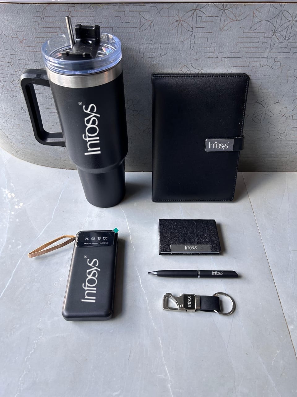 Collection of black branded items including a tumbler, phone case, cardholder, pen, keychain, and wallet on a light gray surface.