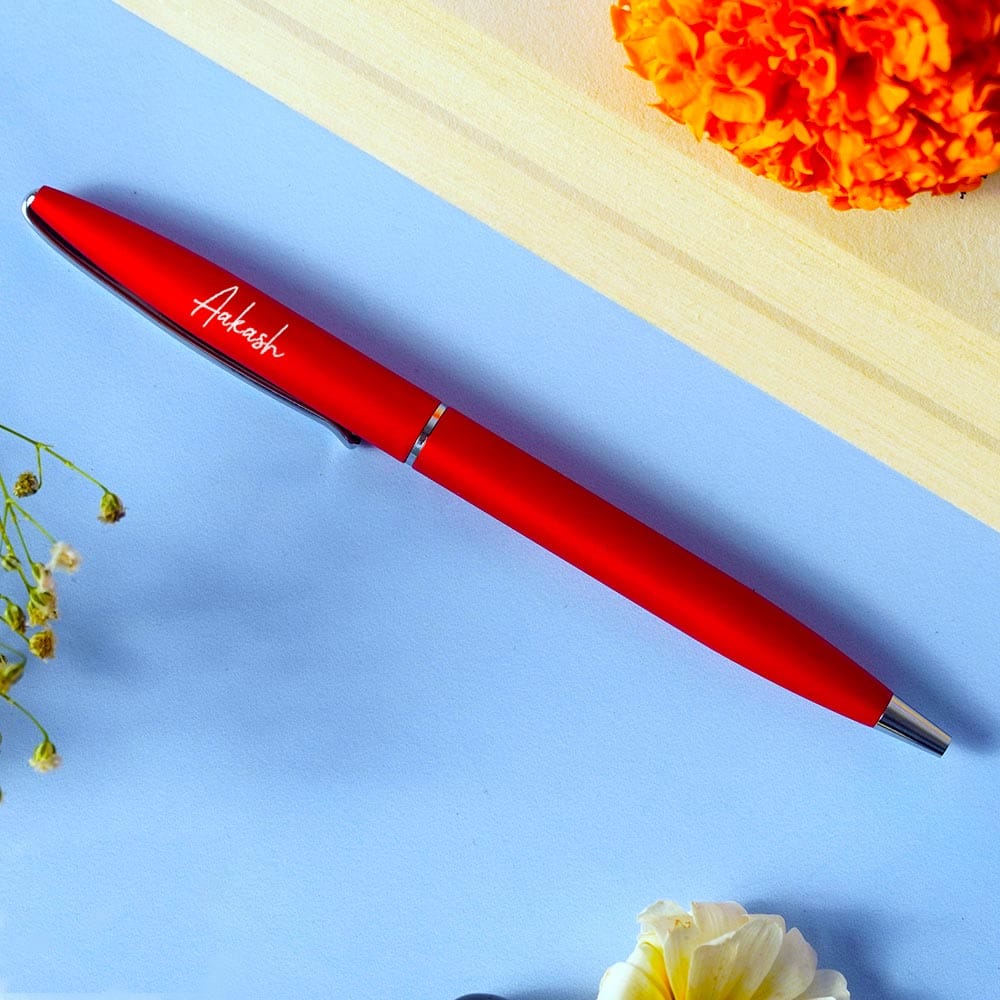 Red pen with 'Ankush' engraved on it, placed on a blue surface with flowers around.