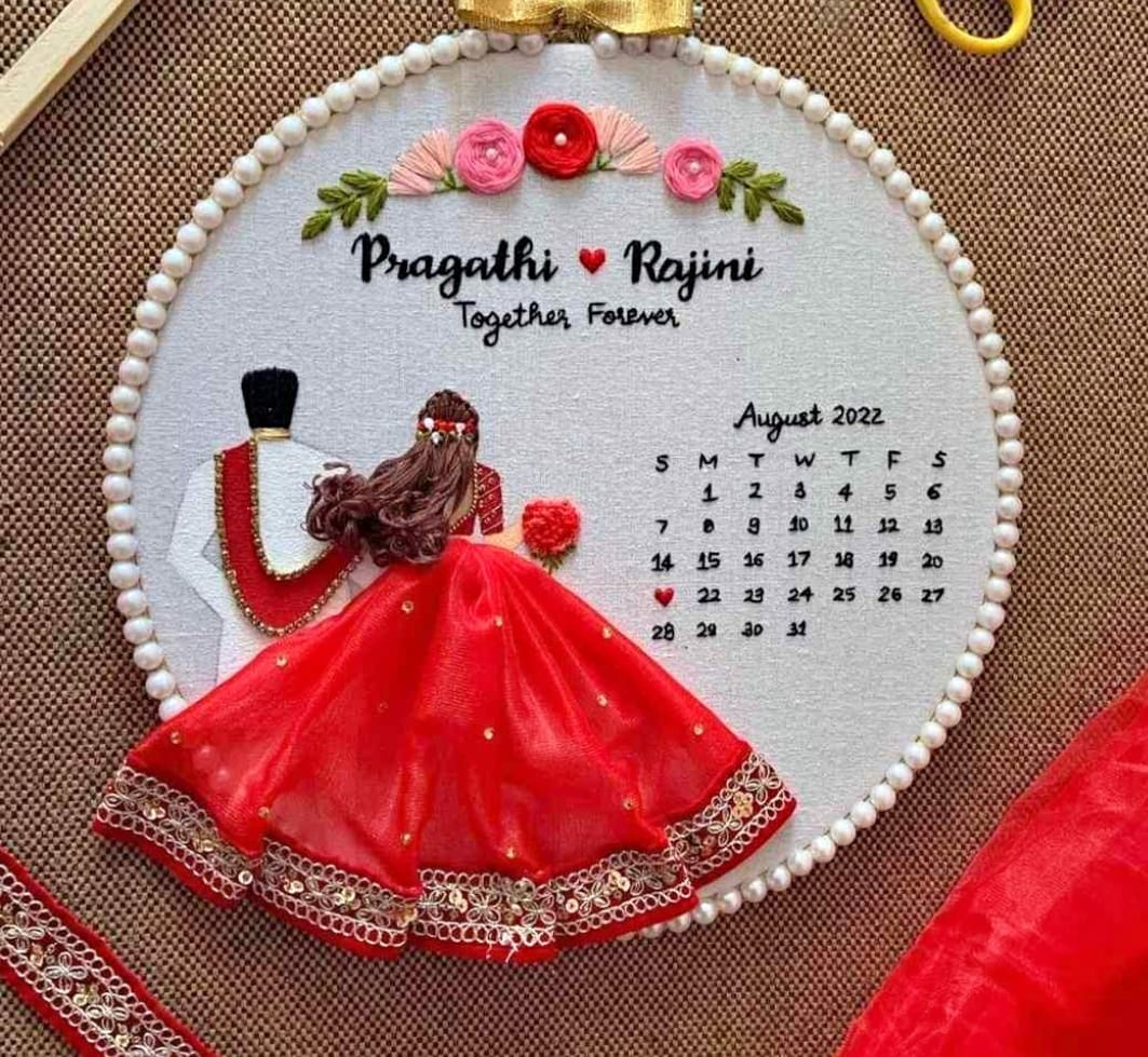 Decorative calendar with embroidered design of a couple and floral elements, featuring the name 'Pragathi Rajini' and the month 'August 2022'.