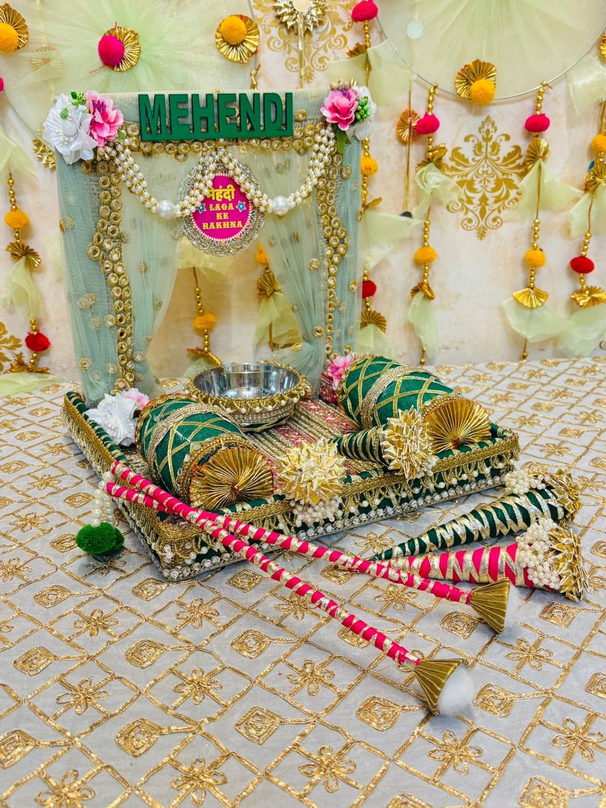 Decorative setup for a Mehendi ceremony with traditional items on a patterned fabric background.