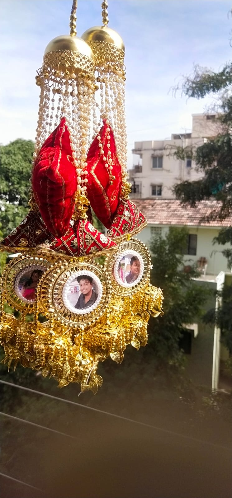 Decorative car hanging with red elements and gold accents, featuring small portraits, against a blurred outdoor background.