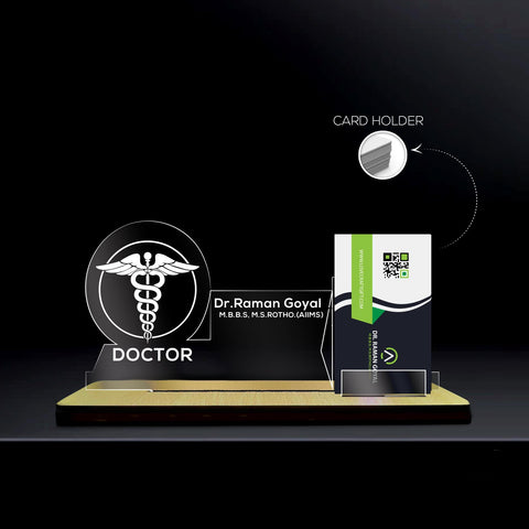 fully customized card holder stand ,A transparent acrylic nameplate with a wooden base, featuring the medical caduceus symbol and the word 'DOCTOR' underneath it. The nameplate includes the name 'Dr. Raman Goyal' with credentials 'M.B.B.S, M.S. RORTHO. (AIIMS)' displayed next to the symbol. On the right side, there is a card holder containing a business card with a QR code and the website 'www.drgsepti.com