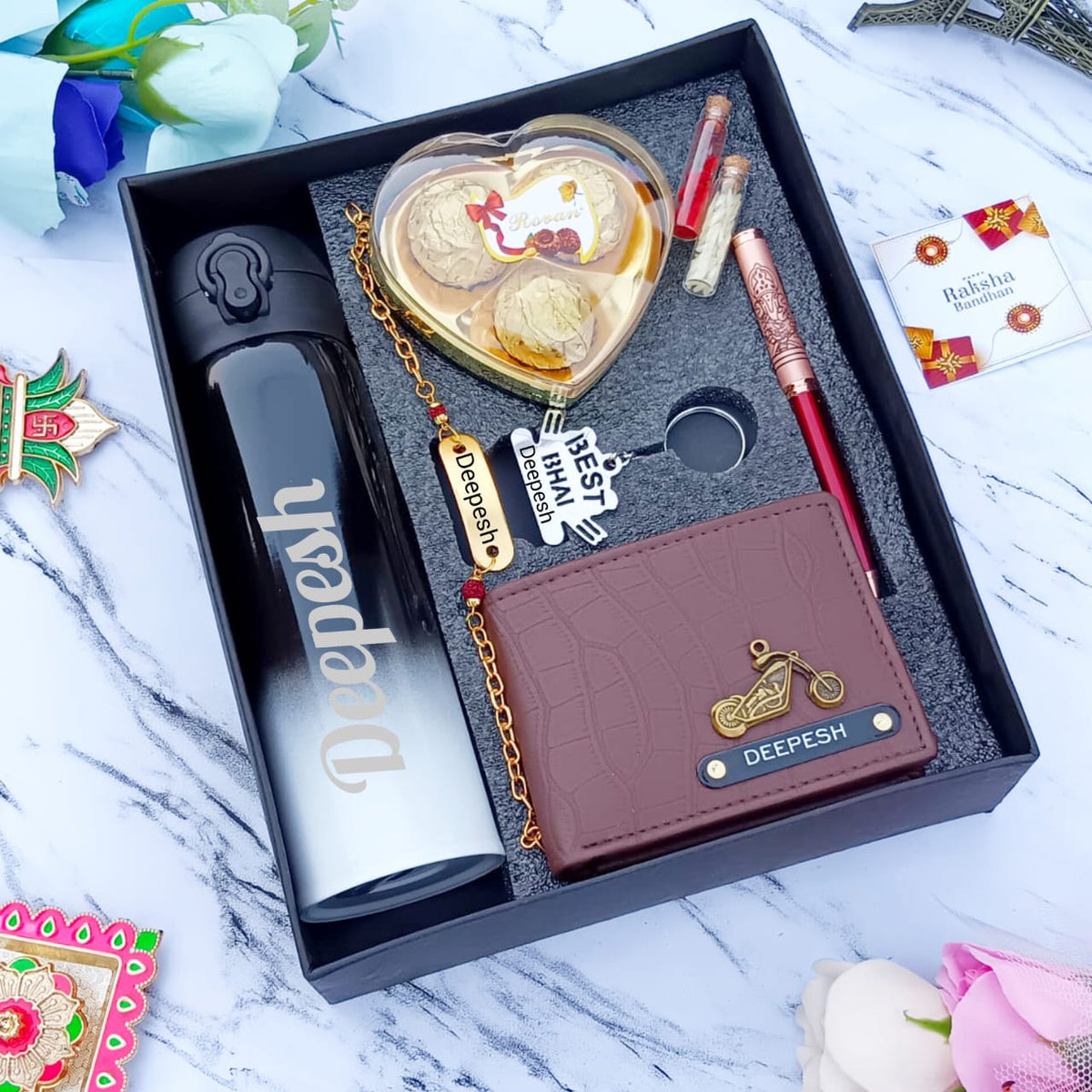 in a black box there one custom flask , chain with brass base on top custom name,heart shape chocolate box,red zari pen,wallet with custom charm and name, 2 small bottles with chawal and roli one greeting card