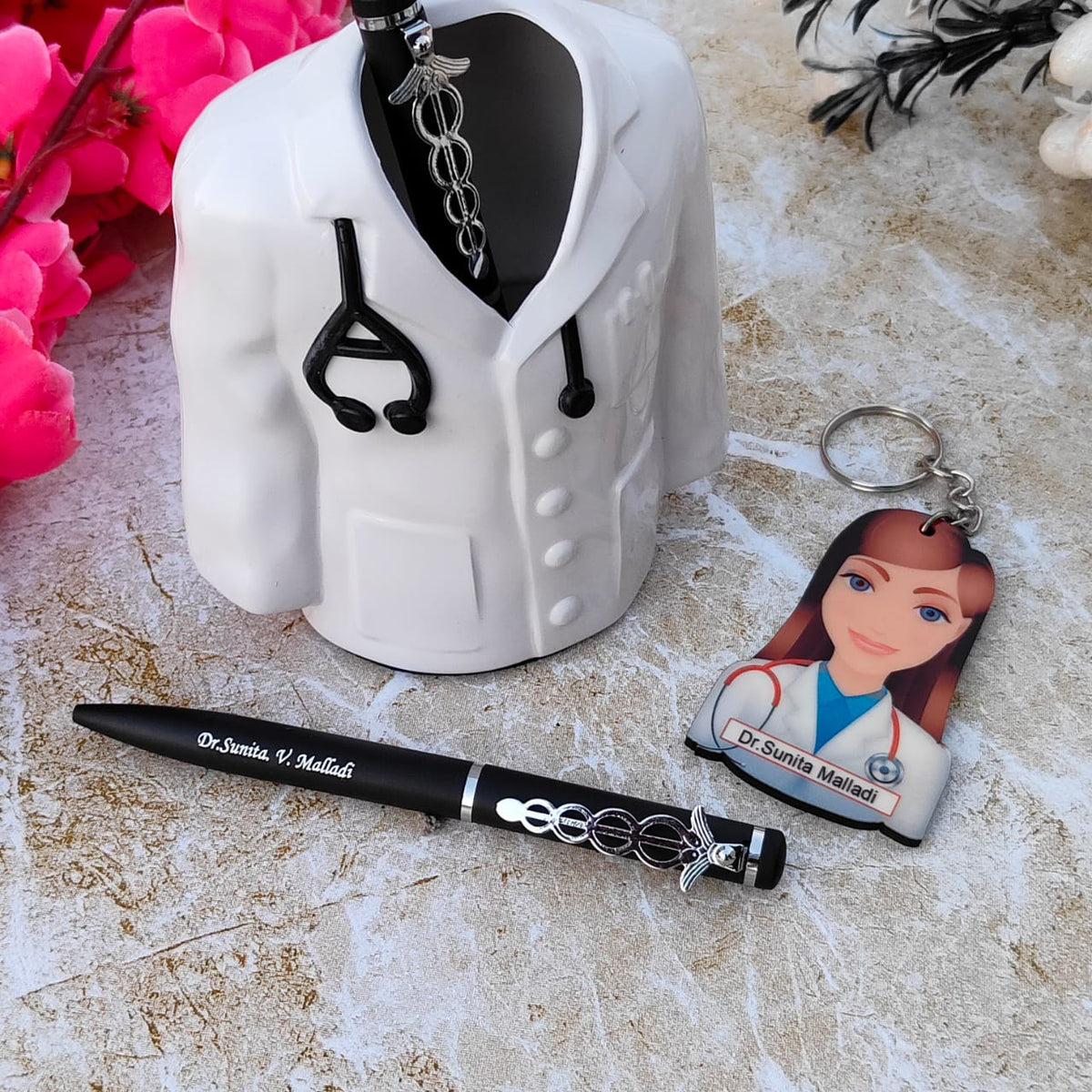 Cooperative gift for doctor A white ceramic pen holder shaped like a doctor's coat with a black stethoscope detail is displayed. A black pen with the custom name engraved on it is placed in the holder, featuring a caduceus charm. Next to the pen holder is a keychain with an illustration of a female doctor and the custom name printed on it. The items are arranged on a light-colored textured surface with pink flowers in the background.