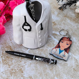 Cooperative gift for doctor A white ceramic pen holder shaped like a doctor's coat with a black stethoscope detail is displayed. A black pen with the custom name engraved on it is placed in the holder, featuring a caduceus charm. Next to the pen holder is a keychain with an illustration of a female doctor and the custom name printed on it. The items are arranged on a light-colored textured surface with pink flowers in the background.