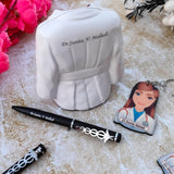 A white ceramic pen holder shaped like a doctor's coat with a  detail is displayed. A black pen with the custom name engraved on it is placed in the holder, featuring a caduceus charm. Next to the pen holder is a keychain with an illustration of a female doctor and the custom name printed on it. The items are arranged on a light-colored textured surface with pink flowers in the background.