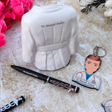 A white ceramic pen holder shaped like a doctor's coat with a detail is displayed. A black pen with the custom name engraved on it is placed in the holder, featuring a caduceus charm. Next to the pen holder is a keychain with an illustration of a male doctor and the custom name printed on it. The items are arranged on a light-colored textured surface with pink flowers in the background.