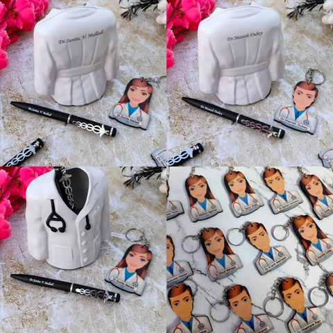 A white ceramic pen holder shaped like a doctor's coat with a black stethoscope detail is displayed. A black pen with the custom name engraved on it is placed in the holder, featuring a caduceus charm. Next to the pen holder is a keychain with an illustration of a female doctor and the custom name printed on it. The items are arranged on a light-colored textured surface with pink flowers in the background.all male female key chains shown in bulk