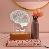 A decorative lamp featuring a detailed wireframe illustration of a human brain, illuminated from a wooden base. The lamp is labeled with the custom title "PSYCHOLOGIST" and the  custom name. It is placed on top of the book.