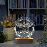 An acrylic LED lamp on a wooden base, featuring the customized name at the top. The lamp displays a detailed illustration of the caduceus symbol with wings, along with a banner reading you can write on your interest   below it. The lamp is placed on a wooden table, with a bouquet of flowers and a books shelf in the background