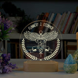 An acrylic LED lamp on a wooden base, featuring the customized name at the top. The lamp displays a detailed illustration of the caduceus symbol with wings, along with a banner reading you can write on your interest   below it. The lamp is placed on a wooden table, with a bouquet of flowers and a vintage books in the background