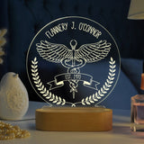 An acrylic LED lamp on a wooden base, featuring the customized name at the top. The lamp displays a detailed illustration of the caduceus symbol with wings, along with a banner reading you can write on your interest   below it. The lamp is placed on a wooden table, with a bouquet of flowers and a vintage stereo in the background