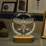 An acrylic LED lamp on a wooden base, featuring the customized name at the top. The lamp displays a detailed illustration of the caduceus symbol with wings, along with a banner reading you can write on your interest   below it. The lamp is placed on a wooden table, with a bouquet of flowers and a vintage stereo in the background