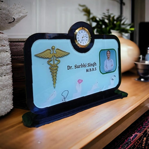 The image shows a personalized desk nameplate for  doctors  The nameplate includes:  A small clock positioned at the top center. A gold medical caduceus symbol on the left side. The custom name and the qualification "M.B.B.S" in the center. A photo of Dr. Surbhi Singh on the right side. Various medical-themed graphics in the background. The nameplate is placed on a wooden surface, and there is a flower pot the background.