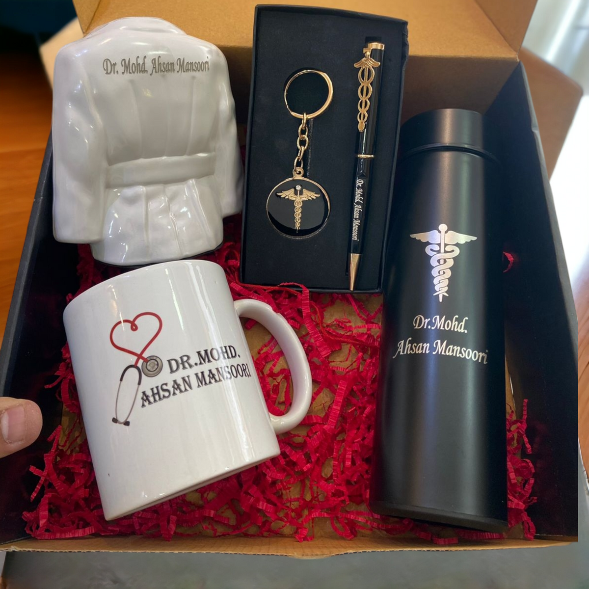 Personalized doctor gift set featuring a balck 500ml temparature bottle with name 'DR.MOHD AHSAN MANSOORI' engraving, a keychain and pen with medical sign, a ceramic doctor coat-shaped pen holder, and a white coffee mug with a red stethoscope heart design and permanent name print. Perfect gift for medical professionals.