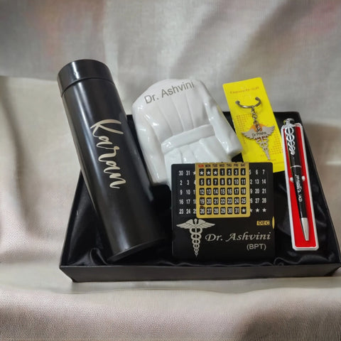 A gift set including a black thermos labeled custom name a pen with a medical symbol, a keychain with a stethoscope charm, a perpetual calendar with custom name inscribed, and a miniature doctor’s coat. The set is presented on a black tray with a pink fabric background.