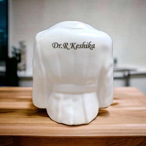 BEST PERSONALIZED CERAMIC  APRON MODEL WHITE PEN STAND with laser permanent print name is "Dr. R Keshika",placed on brown table ,best gifts for doctor, bbdgifts.com