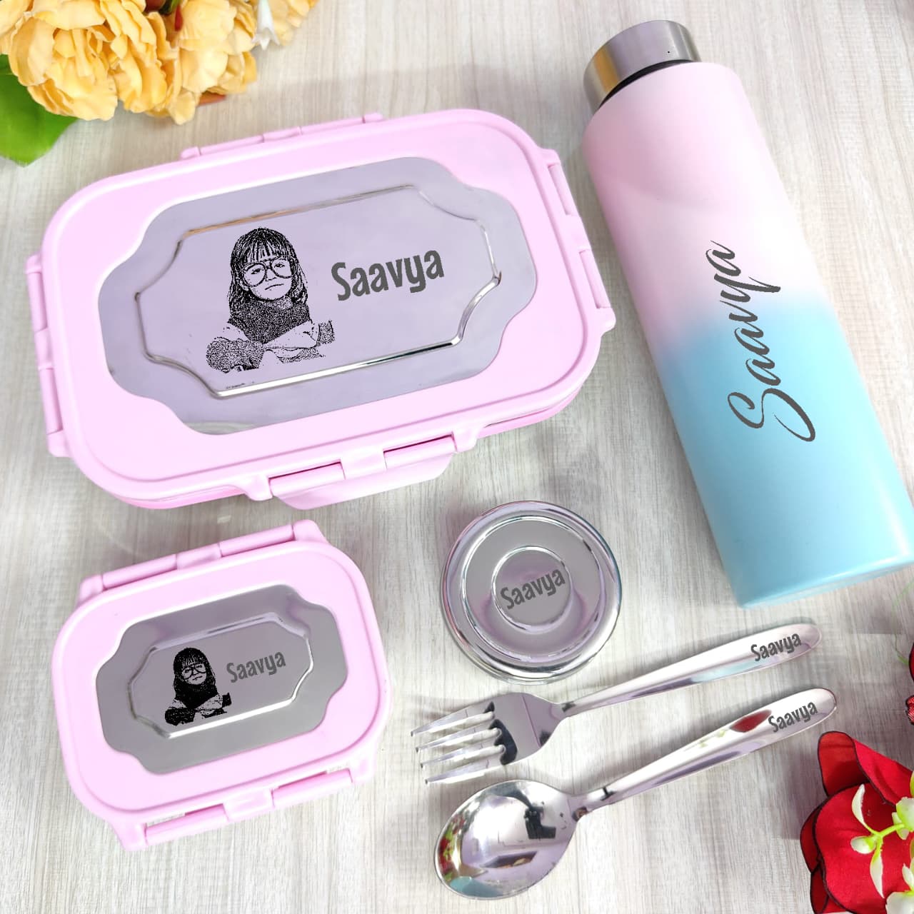 Pink lunch box, water bottle, and cutlery set with 'Saavya' branding on a light wooden surface.