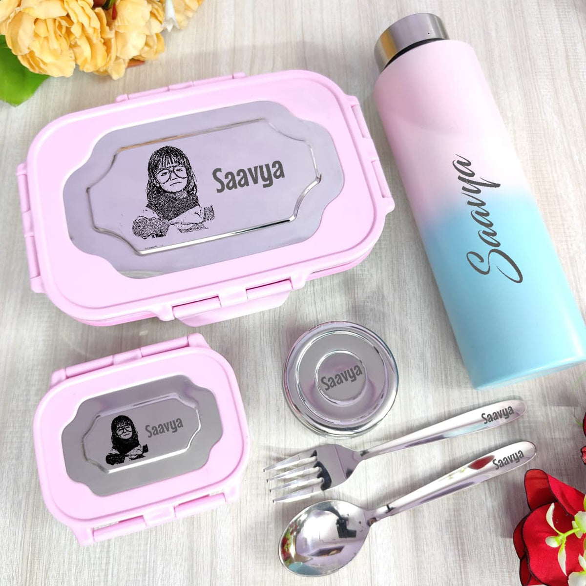 Pink lunch box, water bottle, and cutlery set with 'Saavya' branding on a light wooden surface.