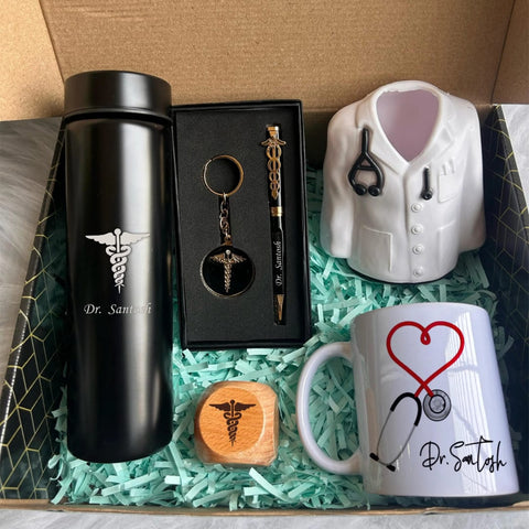 A gift set including a black thermos labeled custom name with a medical symbol, a black pen with a medical symbol and custom name  inscribed, a keychain with a medical symbol, a miniature doctor’s coat, a wooden stamp with a medical symbol, and a white mug with a stethoscope design and custom name inscribed. The set is presented in a box with light blue paper shreds.