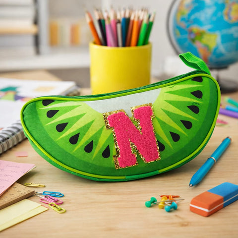 Green pencil case shaped like a watermelon with a pink letter 'N' on a desk with stationery items.