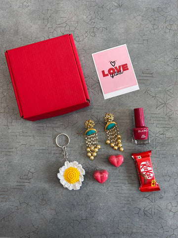 Red gift box with jewelry, a card, a keychain, nail polish, and a candy bar on a textured gray surface.