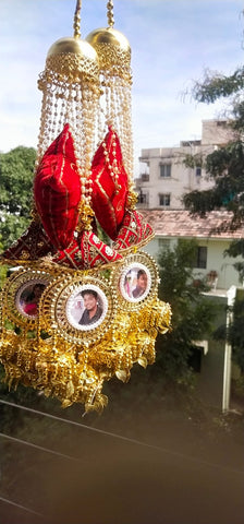 Decorative car hanging with red elements and gold accents, featuring small portraits, against a blurred outdoor background.