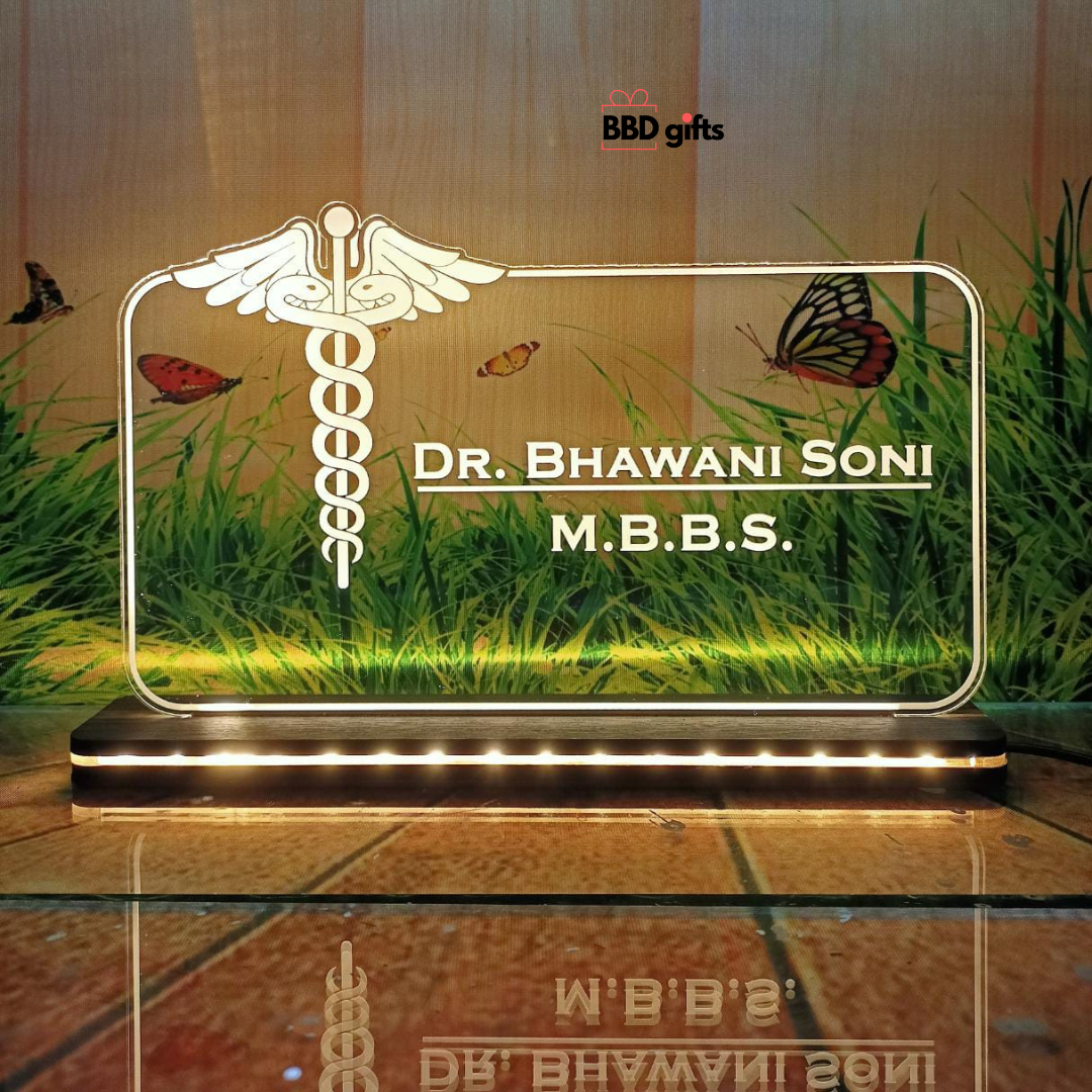 The image shows a personalized desk nameplate for a doctor, featuring a transparent, illuminated glass panel with the medical caduceus symbol. The nameplate reads "Dr. Bhawani Soni, M.B.B.S." and is branded with "BBD gifts" in the background, which depicts grass, butterflies, and a rustic wooden wall.