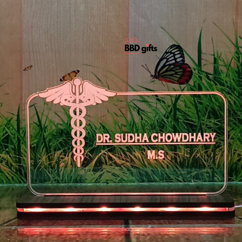 The image shows a personalized desk nameplate for a doctor, featuring a transparent, illuminated glass panel with the medical caduceus symbol. The nameplate reads "Dr. Bhawani Soni, M.B.B.S." and is branded with "BBD gifts" in the background, which depicts grass, butterflies, and a rustic wooden wall. red color light