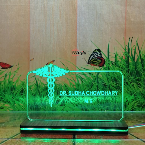 The image shows a personalized desk nameplate for a doctor, featuring a transparent, illuminated glass panel with the medical caduceus symbol. The nameplate reads "Dr. Bhawani Soni, M.B.B.S." and is branded with "BBD gifts" in the background, which depicts grass, butterflies, and a rustic wooden wall. green color light