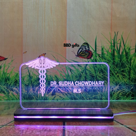 The image shows a personalized desk nameplate for a doctor, featuring a transparent, illuminated glass panel with the medical caduceus symbol. The nameplate reads "Dr. Bhawani Soni, M.B.B.S." and is branded with "BBD gifts" in the background, which depicts grass, butterflies, and a rustic wooden wall. puple color light