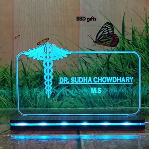 The image shows a personalized desk nameplate for a doctor, featuring a transparent, illuminated glass panel with the medical caduceus symbol. The nameplate reads "Dr. Bhawani Soni, M.B.B.S." and is branded with "BBD gifts" in the background, which depicts grass, butterflies, and a rustic wooden wall. sky blue light color