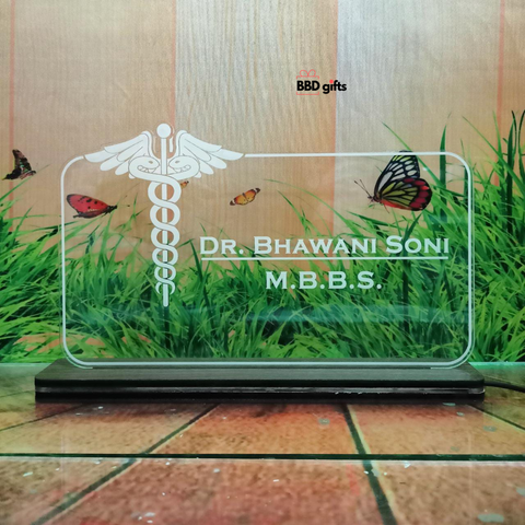 The image shows a personalized desk nameplate for a doctor, featuring a transparent, illuminated glass panel with the medical caduceus symbol. The nameplate reads "Dr. Bhawani Soni, M.B.B.S." and is branded with "BBD gifts" in the background, which depicts grass, butterflies, and a rustic wooden wall. when light is off