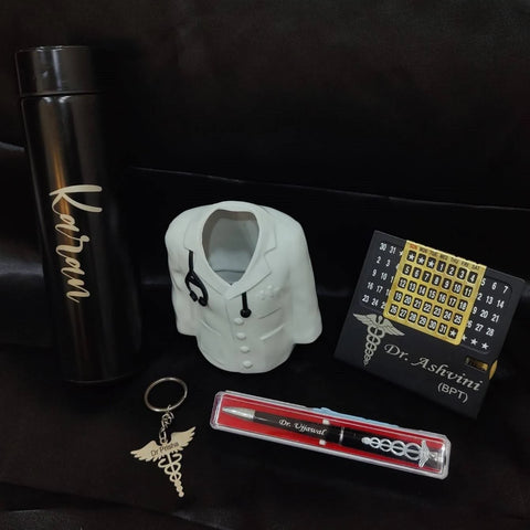 A gift set including a black thermos labeled custom name a pen with a medical symbol, a keychain with a stethoscope charm, a perpetual calendar with custom name inscribed, and a miniature doctor’s coat. The set is placed on floor